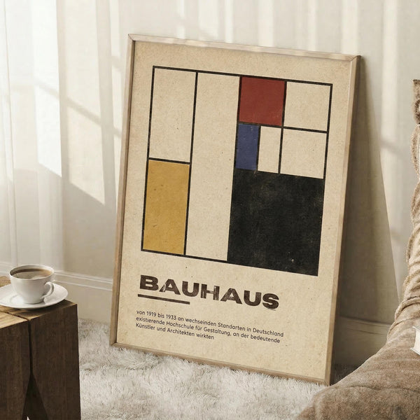 May include: Framed Bauhaus-style geometric poster with colorful rectangles in yellow, red, blue, and black on cream background, leaning against white wall corner next to coffee cup on wooden block and textured throw blanket on white fluffy rug.