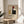 May include: Framed Bauhaus poster with geometric abstract design featuring colored rectangles in red, blue, yellow, and black on cream background, displayed on round wooden side table next to beige upholstered chair with plant leaves visible in corner, natural lighting creating soft shadows on neutral wall.