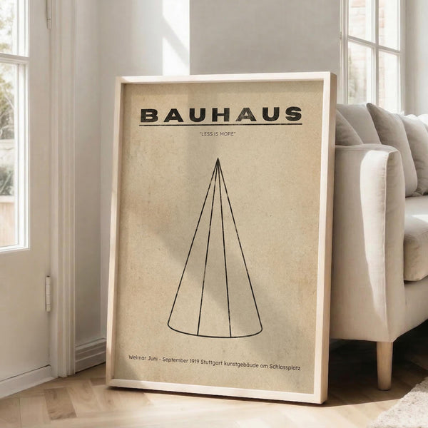 May include: Minimalist Bauhaus poster in beige wooden frame leaning against white wall next to cream sofa. Features geometric cone design with "LESS IS MORE" motto on vintage beige background. German text references Weimar and Stuttgart 1919. Natural lighting from windows creates soft shadows on light wooden floor.