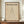 May include: Framed Bauhaus poster with cream background featuring bold black text "BAUHAUS" and "LESS IS MORE" above a minimalist line drawing of a cone shape. German text at bottom references Weimar and Stuttgart 1919. Wooden frame displayed on wood console with white ceramic vase and books in elegant room setting with white walls and trim.