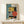 May include: Framed Bauhaus-style geometric poster with abstract architectural shapes in teal, mustard yellow, coral red, and black on beige background, displayed on light wood console table next to small white wrapped gift with twine and pine sprig against cream wall