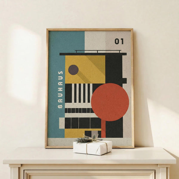 May include: Framed Bauhaus-style geometric poster with abstract architectural shapes in teal, mustard yellow, coral red, and black on beige background, displayed on light wood console table next to small white wrapped gift with twine and pine sprig against cream wall