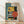 May include: Framed Bauhaus-style geometric poster with abstract architectural elements in yellow, teal, orange, and black colors, featuring circles, rectangles, and linear patterns. The print displays "01" and "BAUHAUS" text with "FORM FOLLOWS FUNCTION" at bottom. Natural wood frame sits on light wooden floor against white wall with soft shadows.