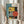May include: Person in cream blouse holding framed geometric poster with abstract architectural forms in teal, yellow, coral, and black colors. Bauhaus-inspired design features circles, rectangles, and linear elements with "01" and "FORM FOLLOWS FUNCTION" text. Natural wood frame, pampas grass visible, neutral home interior setting.