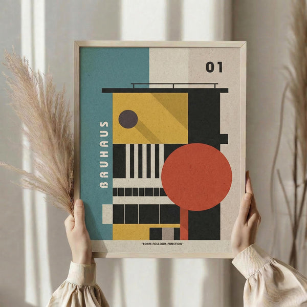 May include: Person in cream blouse holding framed geometric poster with abstract architectural forms in teal, yellow, coral, and black colors. Bauhaus-inspired design features circles, rectangles, and linear elements with "01" and "FORM FOLLOWS FUNCTION" text. Natural wood frame, pampas grass visible, neutral home interior setting.