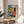 May include: Framed geometric abstract poster in Bauhaus style featuring teal, yellow, orange and black shapes including circles, rectangles and stripes, with "01" numbering and "BAUHAUS" text vertically aligned. Wooden frame sits on round wooden side table next to beige upholstered chair with plant leaves visible, natural sunlight creating soft shadows on white wall.