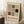 May include: Framed Bauhaus poster with geometric design featuring black, red, and gray squares and rectangles on cream background, displayed in light wood frame leaning against white wall corner. Scene shows white coffee cup on wooden block and beige textured throw blanket on white fuzzy rug.