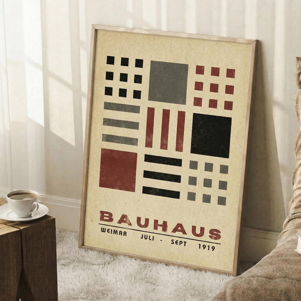 May include: Framed Bauhaus poster with geometric design featuring black, red, and gray squares and rectangles on cream background, displayed in light wood frame leaning against white wall corner. Scene shows white coffee cup on wooden block and beige textured throw blanket on white fuzzy rug.