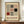 May include: Framed Bauhaus-style geometric poster with black, red, and gray rectangles and squares arranged in a grid pattern on cream background, displayed on wooden sideboard next to white ceramic vase and stacked books in elegant room with white paneled walls