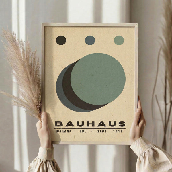 May include: Person holding framed Bauhaus-style poster with geometric design featuring overlapping circles in sage green and dark green on cream background, three small circles above, black text below. Light wood frame, neutral room setting with dried pampas grass and soft natural lighting.