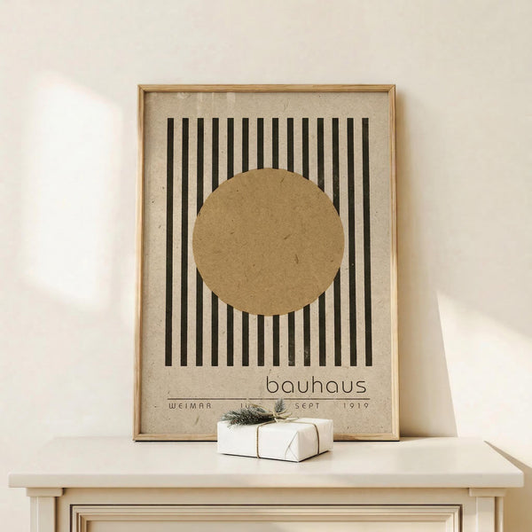 May include: Framed Bauhaus-style poster with beige background, black vertical stripes, and brown circle design, displayed on white wooden surface next to small wrapped gift with evergreen sprig. Warm neutral tones throughout minimalist room setting.