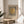 May include: Framed Bauhaus-style poster with vertical black stripes and golden yellow circle, displayed on light wood round side table next to beige upholstered chair. Natural sunlight streams through white curtains, casting soft shadows. Green plant leaves visible in corner. Minimalist modern interior with neutral cream walls and natural textures.