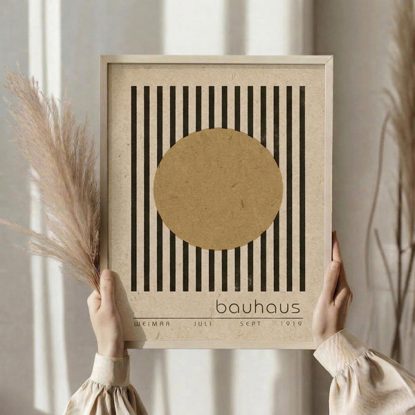 May include: Person in beige clothing holding a framed Bauhaus-style poster featuring vertical black lines with a tan circle overlay on cream background. Dried pampas grass visible to the left. Natural lighting creates soft shadows on neutral wall backdrop.