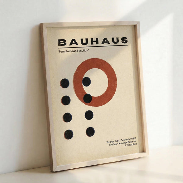 May include: Framed vintage Bauhaus poster with cream background featuring bold black text, red-orange circle, and eight black dots arranged in geometric pattern. Light wood frame leans against white wall, showing minimalist modernist design with German text referencing Weimar 1919 and Stuttgart locations.