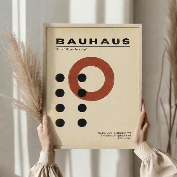 May include: Person holding framed Bauhaus poster with geometric design featuring rust red circle and black dots on cream background, displayed next to dried pampas grass against neutral curtained window setting