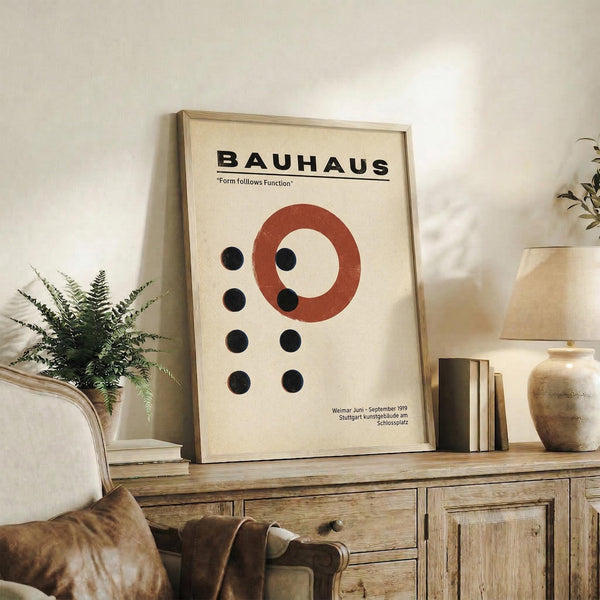 May include: Framed Bauhaus poster with cream background featuring black text, red circular design, and black dots arranged in geometric pattern. Displayed on rustic wooden sideboard with books, ceramic vase, and fern plant. Neutral living room setting with beige chair and brown throw pillows visible.