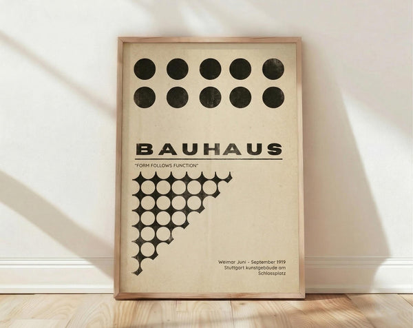 May include: Framed Bauhaus poster with cream background featuring two rows of dark green circles at top, bold black "BAUHAUS" text with "Form Follows Function" subtitle, and geometric pattern of white circles forming triangular shape on black background. Natural wood frame displayed on light wooden floor against white wall with soft shadows.