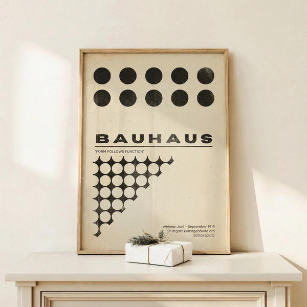May include: Framed Bauhaus poster with geometric black and white circles pattern and "Form Follows Function" text, displayed on light wood surface next to small wrapped gift with twine and greenery sprig, against cream wall with natural lighting