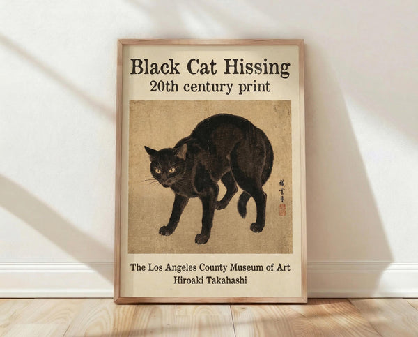 May include: Framed vintage-style poster leaning against white wall on light wood floor. Shows black cat in defensive arched pose with yellow eyes on aged beige background. Includes museum attribution text and Japanese characters. Natural wood frame with warm lighting creating soft shadows.