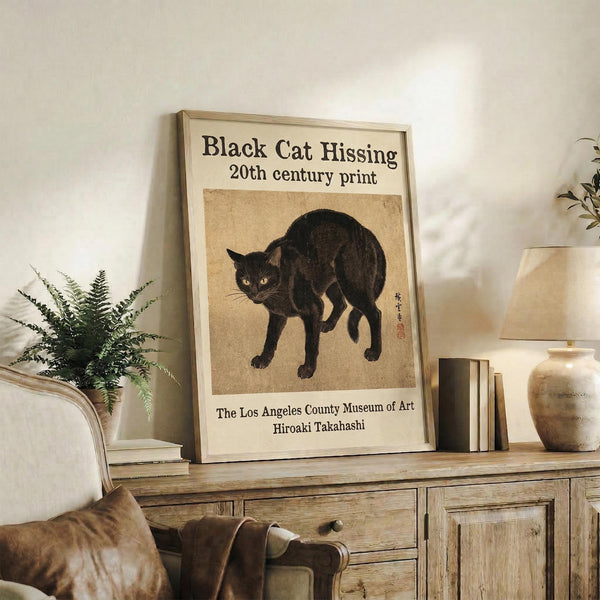 May include: Framed vintage-style poster featuring a black cat with arched back in defensive pose on beige background, displayed on rustic wooden sideboard alongside fern plant, books, and ceramic vase in neutral-toned living room setting with cream upholstered chair visible.