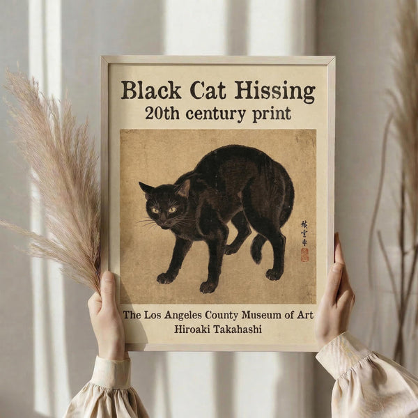 May include: Person holding framed vintage-style poster featuring black cat in defensive hissing pose against aged beige background. Print shows museum attribution text and Japanese characters with red seal. Neutral beige frame matches cream-colored wall setting. Person wears light knit sweater and holds dried pampas grass stems, creating minimalist aesthetic room display.