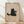 May include: Framed poster print leaning against white wall on wooden floor with natural light shadows. Features cute black cat illustration holding gray coffee mug with pink marshmallows on beige textured background. Word "Meow" appears below cat. Light wood frame, minimalist room setting with white baseboard trim.
