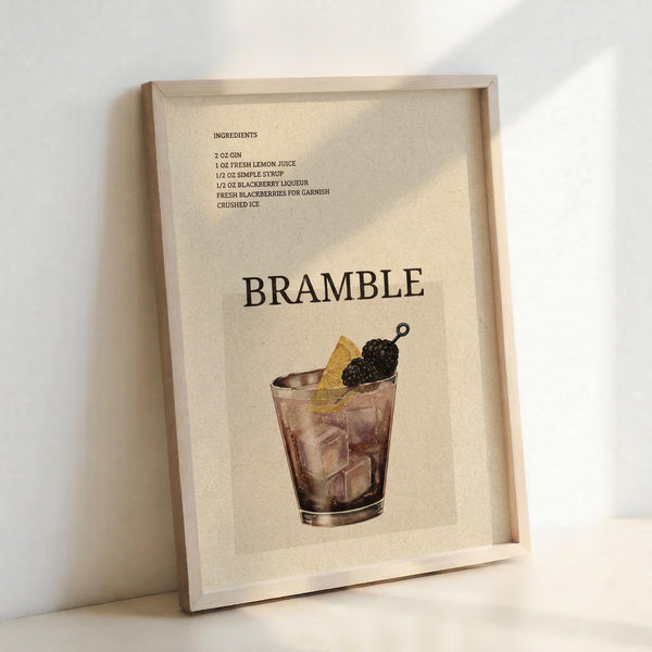May include: Framed poster print leaning against white wall showing vintage-style cocktail recipe for Bramble drink. Cream-colored background with black text listing ingredients including gin, lemon juice, simple syrup, and blackberry liqueur. Features illustrated rocks glass with ice cubes, garnished with fresh blackberries on cocktail pick and lemon slice. Natural wood frame with warm lighting casting soft shadows.