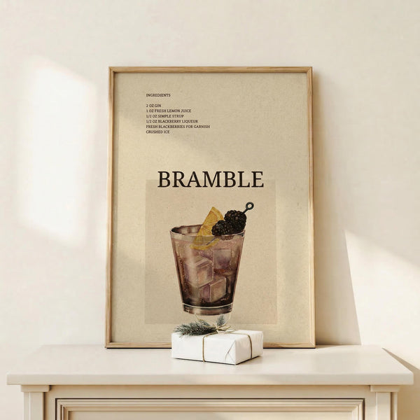 May include: Framed vintage-style cocktail recipe poster featuring a Bramble drink illustration in muted tones, displayed on light wood furniture beside a small wrapped white gift box with evergreen sprig. The beige poster shows ingredients list and glass with blackberry garnish in watercolor style within natural wood frame.
