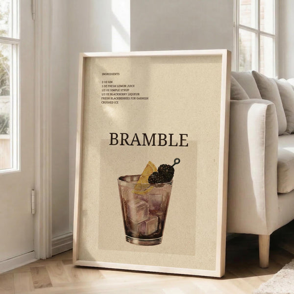 May include: Framed vintage-style cocktail recipe poster displaying "BRAMBLE" in large text with ingredients list above and illustration of purple cocktail in glass with ice, lemon slice and blackberry garnish. Cream-colored background with beige wooden frame, leaning against white wall next to beige upholstered chair in bright room with windows.
