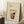 May include: Framed beige poster with illustrated bramble cocktail recipe displayed in cozy living room setting with white walls, wooden stool holding coffee cup, textured rug, and neutral furniture. Artwork shows dark cocktail glass with blackberry garnish and ingredient list in brown text against cream background.