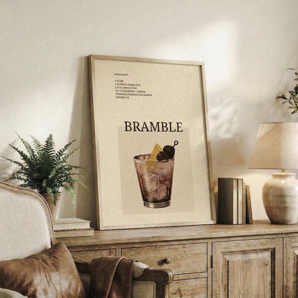 May include: Framed beige poster displaying bramble cocktail recipe with ingredients list at top and large "BRAMBLE" text above illustrated glass filled with dark liquid, ice cubes, and blackberry garnish. Poster leans against white wall on rustic wooden sideboard next to fern plant, books, ceramic vases, and brown leather chair in warm-toned living room setting.