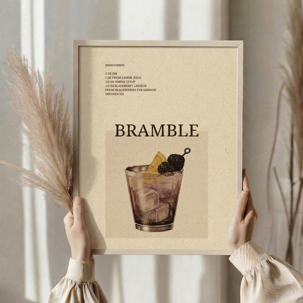 May include: Hands holding a framed cocktail recipe poster featuring a Bramble drink illustration with blackberry garnish. The beige-toned print shows ingredients list and is displayed in a light wood frame. Person wears cream-colored ruffled sleeves while holding dried pampas grass stems. Soft neutral interior setting with white curtains in background.