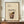 May include: Framed vintage-style cocktail recipe poster displaying a Bramble drink illustration with ingredients list, shown in brown wooden frame on wooden credenza beside white ceramic vase and books against cream paneled wall with decorative brass sconce