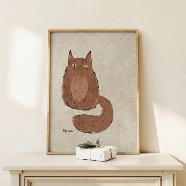 May include: Framed artwork showing a fluffy brown cat with yellow eyes and pointed ears sitting with its long tail curled around, with "Meow" written below in brown text on a beige textured background. The wooden frame sits on a light wood surface next to a small white gift box with evergreen sprig, against a cream wall with soft natural lighting.