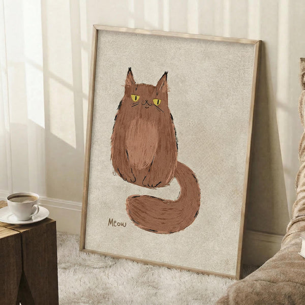 May include: Framed artwork featuring a fluffy brown cat with bright yellow eyes sitting upright with curved tail, painted in loose brushstroke style on cream textured background with "Meow" text below. Light wood frame leaning against white wall near white coffee cup on wooden block and beige textured fabric in soft natural lighting.