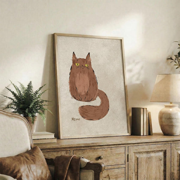 May include: Framed artwork featuring a fluffy brown cat with yellow eyes and pointed ears sitting upright with its long tail curled around, with "Meow" text below, displayed on a rustic wooden sideboard alongside books, ceramic vases, a fern plant, and cream-colored furniture in a cozy living room setting with neutral walls and warm lighting.