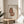 May include: Framed poster print featuring a fluffy brown cat with bright yellow-green eyes and long bushy tail, sitting upright with "Meow" text below. The artwork has a hand-drawn watercolor style on cream textured background in a thin wooden frame. Displayed on a round light wood side table next to a beige upholstered chair and plant leaves, with natural sunlight streaming across neutral walls.
