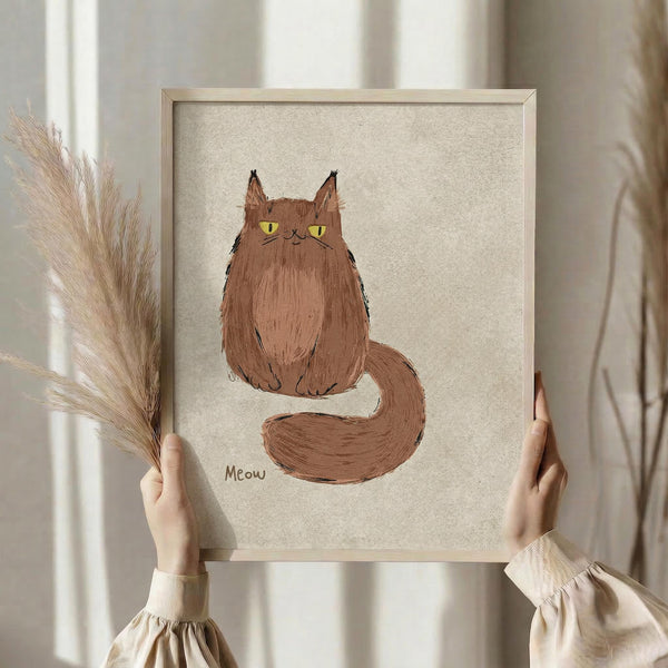 May include: Person holding framed artwork showing a fluffy brown cat with yellow eyes and long tail sitting upright, with "Meow" text below. Light wood frame on textured beige background. Person wearing cream ruffle-sleeve top holds dried pampas grass stems. Soft natural lighting by window with white curtains.