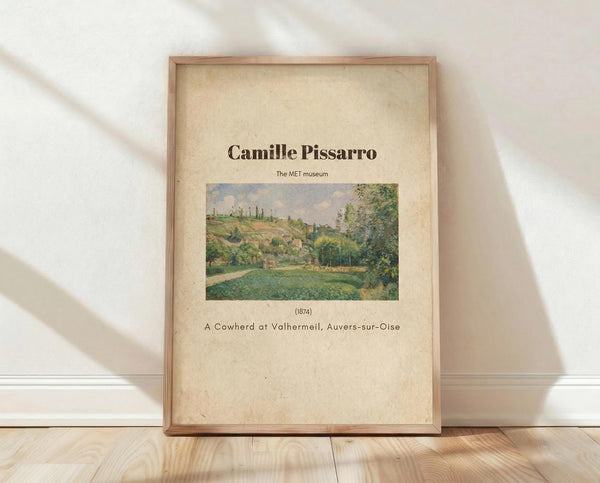 May include: Framed art print featuring Camille Pissarro's 1874 painting of a pastoral countryside scene with green fields, trees, and rolling hills under a cloudy sky. The beige poster displays "Camille Pissarro" at the top, "The MET museum" below, and "A Cowherd at Valhermeil, Auvers-sur-Oise (1874)" at the bottom. Natural wood frame leans against white wall on light wooden floor with soft shadows.