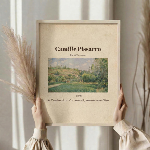May include: hands holding a framed art print featuring a pastoral landscape painting with green fields, trees, and rolling hills in impressionist style. The beige matted frame displays text identifying the artist and artwork. Dried pampas grass visible to the left, neutral beige clothing and soft natural lighting create a minimalist aesthetic against white curtained background.