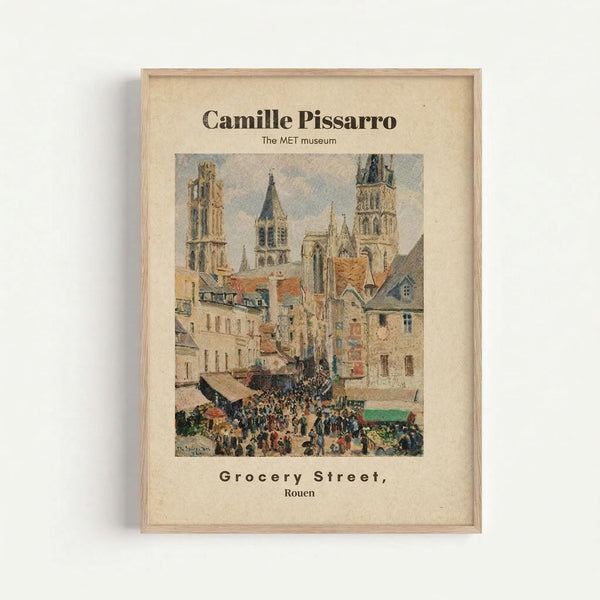 Framed reproduction of a Pissarro painting showing a bustling French street scene in Rouen with Gothic cathedral towers, colorful medieval buildings, and crowds of people below. The artwork features muted earth tones and impressionist brushwork, displayed in a simple light wood frame against a white wall. Text above reads "Camille Pissarro The MET museum" and below "Grocery Street, Rouen."