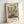 May include: Framed poster print displaying Camille Pissarro's impressionist painting of a bustling French street scene in Rouen. The artwork shows medieval cathedral towers, colorful buildings with orange roofs, and crowds of people in a market square. Text at top reads "Camille Pissarro The MET museum" and bottom shows "Grocery Street, Rouen". Light wooden frame against white wall with natural lighting casting soft shadows.