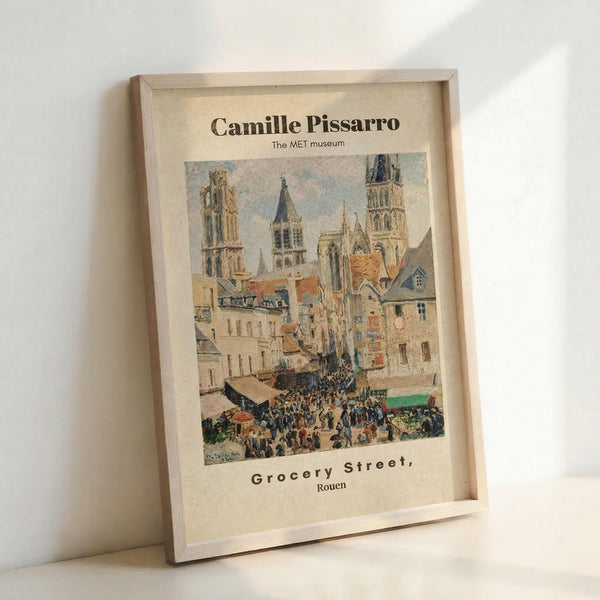 May include: Framed poster print displaying Camille Pissarro's impressionist painting of a bustling French street scene in Rouen. The artwork shows medieval cathedral towers, colorful buildings with orange roofs, and crowds of people in a market square. Text at top reads "Camille Pissarro The MET museum" and bottom shows "Grocery Street, Rouen". Light wooden frame against white wall with natural lighting casting soft shadows.