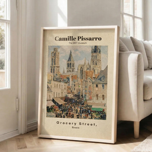 May include: Framed museum poster print leaning against white wall next to beige sectional sofa in bright living room. The vintage-style artwork shows a busy European street scene with Gothic cathedral towers, colorful buildings, and crowds of people. Cream matting surrounds the impressionist painting reproduction in light wood frame on hardwood floors with area rug.