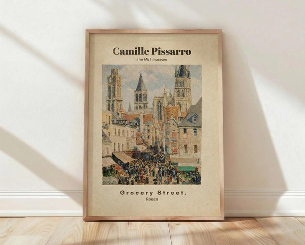 May include: Framed art print leaning against white wall on light wood floor. Shows Impressionist painting of bustling French street scene with Gothic cathedral towers, colorful buildings with orange roofs, and crowds of people below. Warm beige matting in natural wood frame. Soft natural lighting creates gentle shadows on textured white wall.