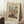 May include: Framed Camille Pissarro reproduction poster leaning against white wall on fluffy white rug. Print shows bustling French street scene in Rouen with Gothic cathedral towers, colorful buildings, and crowds of people. Warm beige mat surrounds the impressionist artwork in natural wood frame. Coffee cup on wooden block and cozy blanket visible nearby, creating peaceful home setting with soft natural lighting.