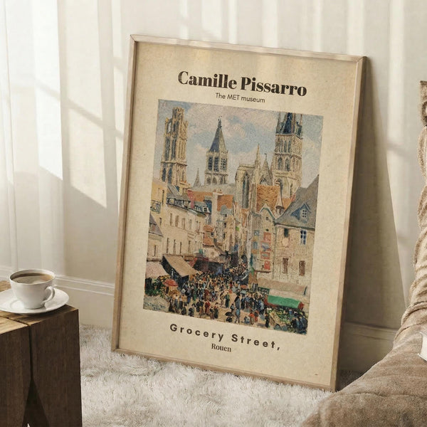 May include: Framed Camille Pissarro reproduction poster leaning against white wall on fluffy white rug. Print shows bustling French street scene in Rouen with Gothic cathedral towers, colorful buildings, and crowds of people. Warm beige mat surrounds the impressionist artwork in natural wood frame. Coffee cup on wooden block and cozy blanket visible nearby, creating peaceful home setting with soft natural lighting.