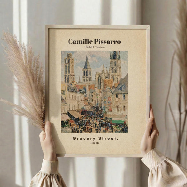 May include: Hands holding a framed art print featuring Camille Pissarro's painting of a bustling French street scene with Gothic cathedral towers, market crowds, and colorful buildings. The beige matted print shows "The MET museum" and "Grocery Street, Rouen" text. Dried pampas grass visible on left side against neutral curtained background.