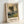 May include: Framed art print of an impressionist landscape painting featuring tall trees along a country path at sunset, with soft golden and green tones throughout the pastoral scene. The wooden frame leans against a white wall, showing the cream-colored matting and black text identifying the artwork and artist below the image.