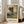 May include: Framed art print of an impressionist landscape painting featuring tall trees along a winding path at sunset, displayed in a bright living room corner next to a beige armchair. The artwork shows green foliage and golden sky in soft, textured brushstrokes within a light wooden frame against white walls and hardwood floors.